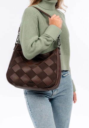 Personne portant un pull en tricot vert clair et un jean bleu, tenant un sac à bandoulière en cuir tressé marron foncé sur le bras.