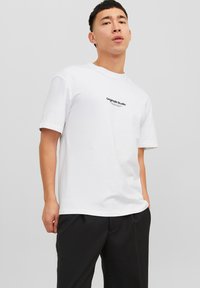 White cotton T-shirt with short sleeves, features "Originals Studio" text in black across the chest. Relaxed fit, smooth texture.