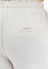 Ljust beige byxor med en mjuk textur, som har ett elastiskt midjeband och en enda bakficka. Enkelt, rent design utan mönster.