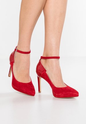 Escarpins rouges en daim et cuir verni, dotés d'une bride cheville, d'un bout pointu et d'un talon aiguille. Texture lisse avec des finitions contrastées.