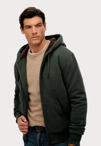 Donkergroene zip-up hoodie met een gestructureerde bruine voering. Heeft twee voorzakken en een relaxte pasvorm over een beige trui.