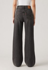 Jean en denim noir à taille haute et jambes larges, avec une texture légèrement délavée, des poches arrière intégrées et un patch en cuir marqué.