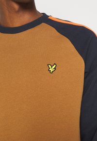 Homme portant un sweat marron avec des manches bleu marine, une rayure orange sur l'épaule et un petit emblème d'oiseau jaune sur la poitrine.