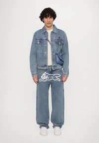 Giacca in denim blu chiaro con due tasche anteriori, abbinata a jeans blu chiaro a vestibilità ampia con un design grafico bianco e sneaker bianche.