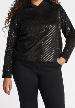 Kobieta ubrana w czarną bluzkę z długim rękawem z diagonalnymi paskami z cekinów i czarne spodnie, jedna ręka w kieszeni, na jednolitym tle.