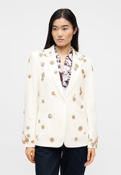 Γυναίκα που φοράει λευκό σακάκι στολισμένο με χρυσές και ασημένιες εφαρμογές λουλουδιών πάνω από ένα εμπριμέ πουκάμισο και σκούρα τζιν, σε απλό φόντο.