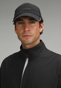 Chaqueta de running negra con cuello alto, cierre de cremallera y tejido texturizado. Gorro negro con logo. La cara del modelo es parcialmente visible, fondo neutro.