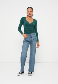 Grünes Langarm-Oberteil mit V-Ausschnitt und geraffter Textur, kombiniert mit hellblauen geraden Jeans und schwarzen Sneakern.