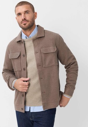 Mand iført brun knappet jakke over beige sweater og lyseblå skjorte, poserer mod en ensfarvet lys baggrund.
