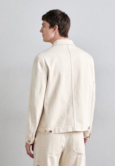 Light beige denim jacket with a relaxed fit, featuring a collared neckline, buttoned cuffs, and gold hardware accents. Simple, clean design.