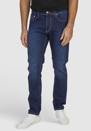 Dark blue denim jeans with a straight leg design, five pockets, and contrast stitching, styled with a black shirt and white sneakers.