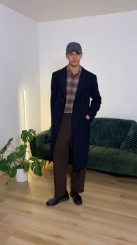 Hosszú sötétkék kabát kockás ing fölött, barna nadrággal és fekete cipővel párosítva. A háttérben zöld kanapé és egy növény látható.