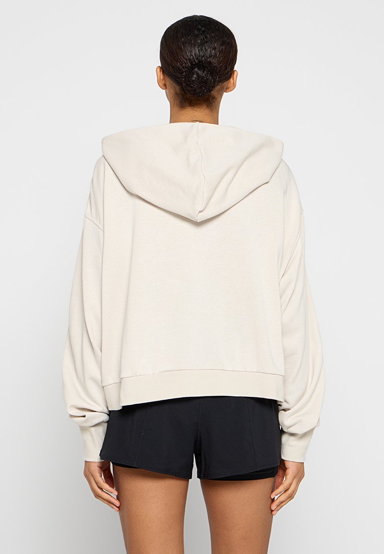 Felpa corta beige con cappuccio regolabile, maniche ampie e orlo a costine, realizzata in tessuto morbido. Indossata sopra pantaloni corti neri.