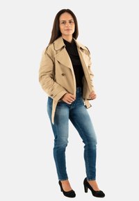 Donna con lunghi capelli castani che indossa un trench beige, una maglietta nera, jeans blu e tacchi neri, in piedi contro uno sfondo chiaro e uniforme.