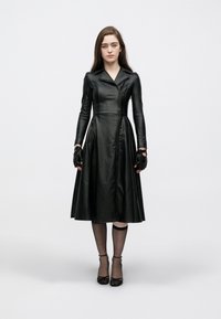 Μαύρο δερμάτινο φόρεμα με εφαρμοστό μπούστο και φούστα που φαρδαίνει, με γιακά, μπροστινό φερμουάρ και πλαϊνές τσέπες. Φοριέται με γάντια χωρίς δάχτυλα και μαύρες γόβες.