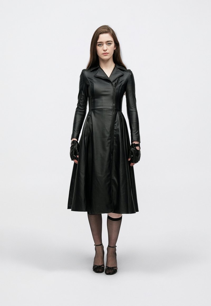 Μαύρο δερμάτινο φόρεμα με εφαρμοστό μπούστο και φούστα που φαρδαίνει, με γιακά, μπροστινό φερμουάρ και πλαϊνές τσέπες. Φοριέται με γάντια χωρίς δάχτυλα και μαύρες γόβες.