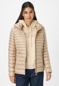 Beige pufferjack met horizontaal gestikt ontwerp, capuchon, ritsen aan de voorkant, zijzakken, gedragen over een lichtbeige hoodie, gecombineerd met blauwe jeans.