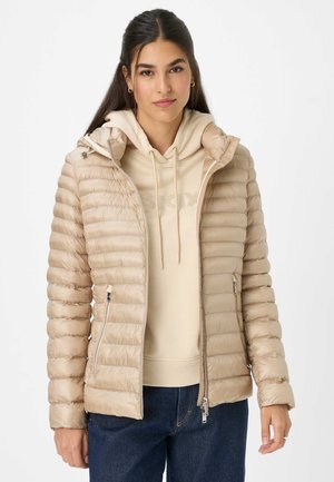 Beige pufferjack met horizontaal gestikt ontwerp, capuchon, ritsen aan de voorkant, zijzakken, gedragen over een lichtbeige hoodie, gecombineerd met blauwe jeans.