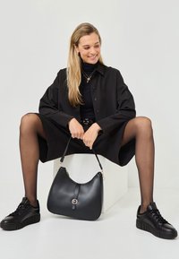 Svart läderhandväska med en böjd silhuett, guldfärgad hårdvara och en slät textur. Modellen bär svarta kläder och sneakers.