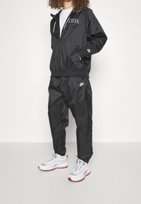 Osoba oblečená v černém sportovním obleku Nike s bílými teniskami s červenými akcenty, stojící před jednobarevným bílým pozadím.
