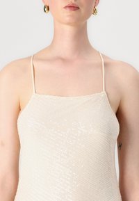 Camisole beige en sequins avec de fines bretelles spaghetti. Présente une surface texturée et brillante avec un décolleté droit, mettant en avant un design minimaliste.