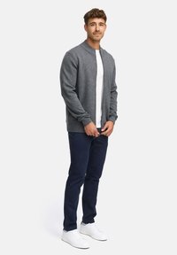 Cardigan gris en tricot avec un col montant, porté sur un t-shirt blanc, associé à un pantalon bleu marine et des baskets blanches.