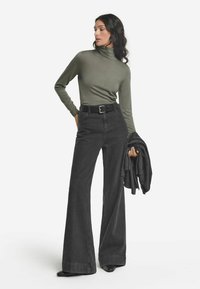 Olivgrön polotröja, matchad med högmidjade svarta utsvängda byxor. Svart bälte med en silverspänne. Modell som håller en mörkgrå jacka.