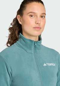 Giacca zip-up in pile di colore blu teal con colletto alto. Presenta una zip frontale e il logo TERREX bianco sul lato sinistro del petto. Texture morbida.