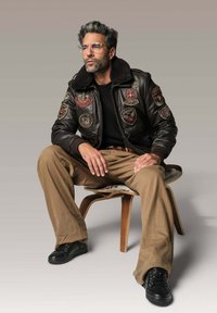 Homme aux cheveux gris et portant des lunettes, assis sur une chaise en bois, vêtu d'une veste en cuir noire avec des écussons, de pantalons beiges et de chaussures noires.