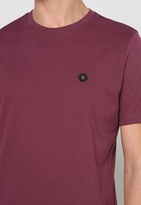 Bordeauxrood katoenen T-shirt met ronde hals, korte mouwen en een klein zwart zeshoekig patch met drie gouden strepen op de borst.