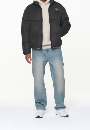 Zwarte pufferjas met een zachte textuur, rits aan de voorkant en geborduurd merklogo, gecombineerd met lichtblauwe jeans met rechte pijpen en witte sneakers.