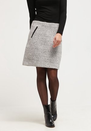 Kvinde iført en knælange grå uldnederdel med en sort lynlåslomme, kombineret med gennemsigtige sorte strømpebukser og sorte ankelstøvler.