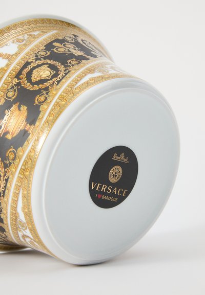 Xícara de cerâmica branca com padrões ornamentais barrocos em preto e dourado e logótipo da Versace na base, inclinada para o lado.