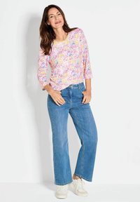 Bloemige top met lange mouwen in roze, gele en blauwe tinten, gecombineerd met lichtblauwe rechttoe rechtaan jeans en witte sneakers.