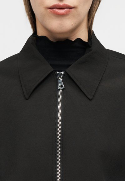 Κοντινό πλάνο ατόμου που φοράει μαύρο μπουφάν με ασημένιο φερμουάρ και μαύρο τοπ με βολάν και ψηλό γιακά από κάτω.