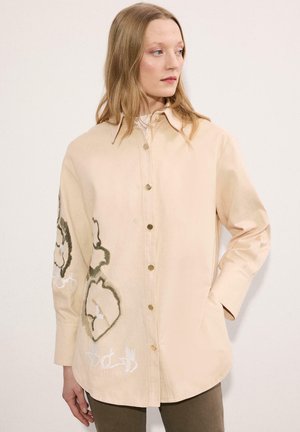 Femeie purtând o cămașă bej cu nasturi, cu broderie florală mare pe partea stângă și pe mânecă, stând pe un fundal alb simplu.