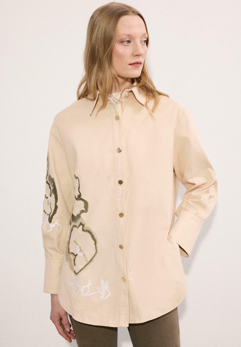 Γυναίκα που φοράει μπεζ πουκάμισο με κουμπιά, με μεγάλη κεντητή άνθη δεξιά στο μπροστινό μέρος και στο μανίκι, στέκεται μπροστά σε απλό λευκό φόντο.