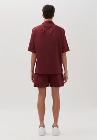 Camicia a maniche corte color marrone bordeaux e pantaloni corti abbinati, realizzati in tessuto liscio; indossati con sneaker bianche; visti da dietro su uno sfondo chiaro.