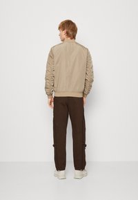 Beige Bomberjacke mit gerafften Ärmeln, gerippten Bündchen und tailliertem Saum, kombiniert mit dunkelbraunen tapered Hosen und hellen Schuhen.