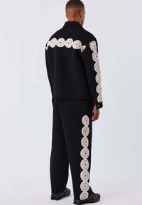 Schwarze Strickjacke und -hose mit großen kreisförmigen cremefarbenen Akzenten an den Seiten. Lockere Passform mit seitlichen Schlitzen an der Jacke.