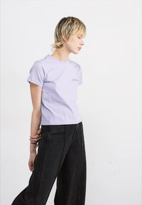 T-shirt viola chiaro a maniche corte con scollo rotondo e piccolo logo sul petto, abbinato a pantaloni neri a vita alta e fondo largo.