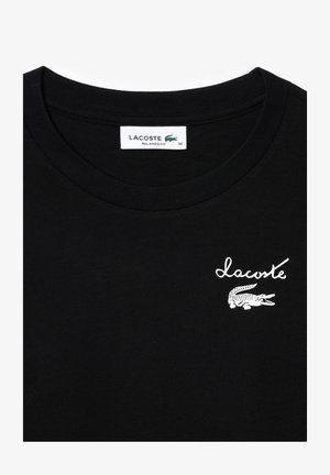 Juodas medvilnės marškinėliai su apvaliu iškirpimu. Priekyje yra baltas "Lacoste" logotipas ir krokodilo grafika. Laisvas kirpimas, dydis 36.