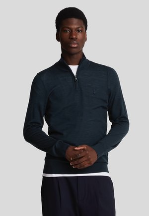 Pull-over en navire sombre avec un col zippé mi-hauteur, des poignets et un ourlet côtelés, arborant un petit logo sur la poitrine gauche. Fabriqué en tissu tricoté lisse.