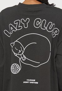 Контур на спяща котка с топка прежда под текста "LAZY CLUB" на тъмна риза, с отпечатано "10:00 AM DON'T DISTURB" под него.