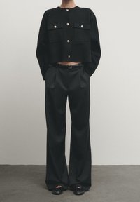 Svart kort jacka med två framfickor och silverknappar, kombinerad med svarta byxor med vida ben och öppna svarta skor.