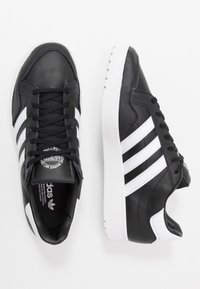 Svarta lädersneakers med vita ränder, med rund tå, snörning framtill och platt vit gummisula. Slät textur och klassisk design.