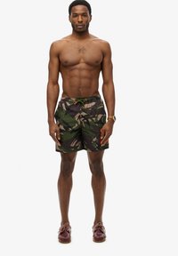 Shorts de bain camouflage en vert, marron et noir, avec un dos élastique, un cordon de serrage orange et des accents de poche contrastants.
