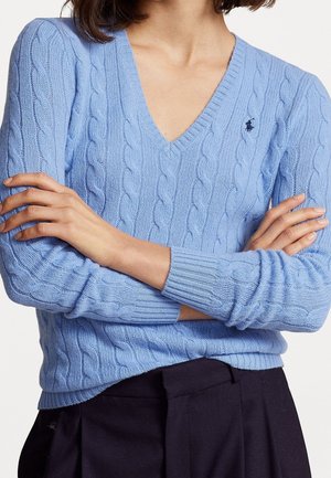Mujer con los brazos cruzados, vistiendo un suéter de punto trenzado azul claro con escote en V y pantalones oscuros, mostrando el torso superior y las manos.