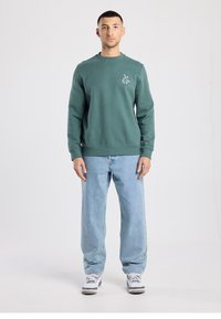 Green sweatshirt made of cotton with ribbed cuffs and hem; features a small embroidered design on the left chest. Paired with blue denim jeans.