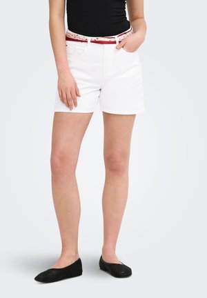 Femme portant un short blanc, un haut noir, une ceinture rouge et blanche et des chaussures plates tissées noires, debout avec une main dans la poche.